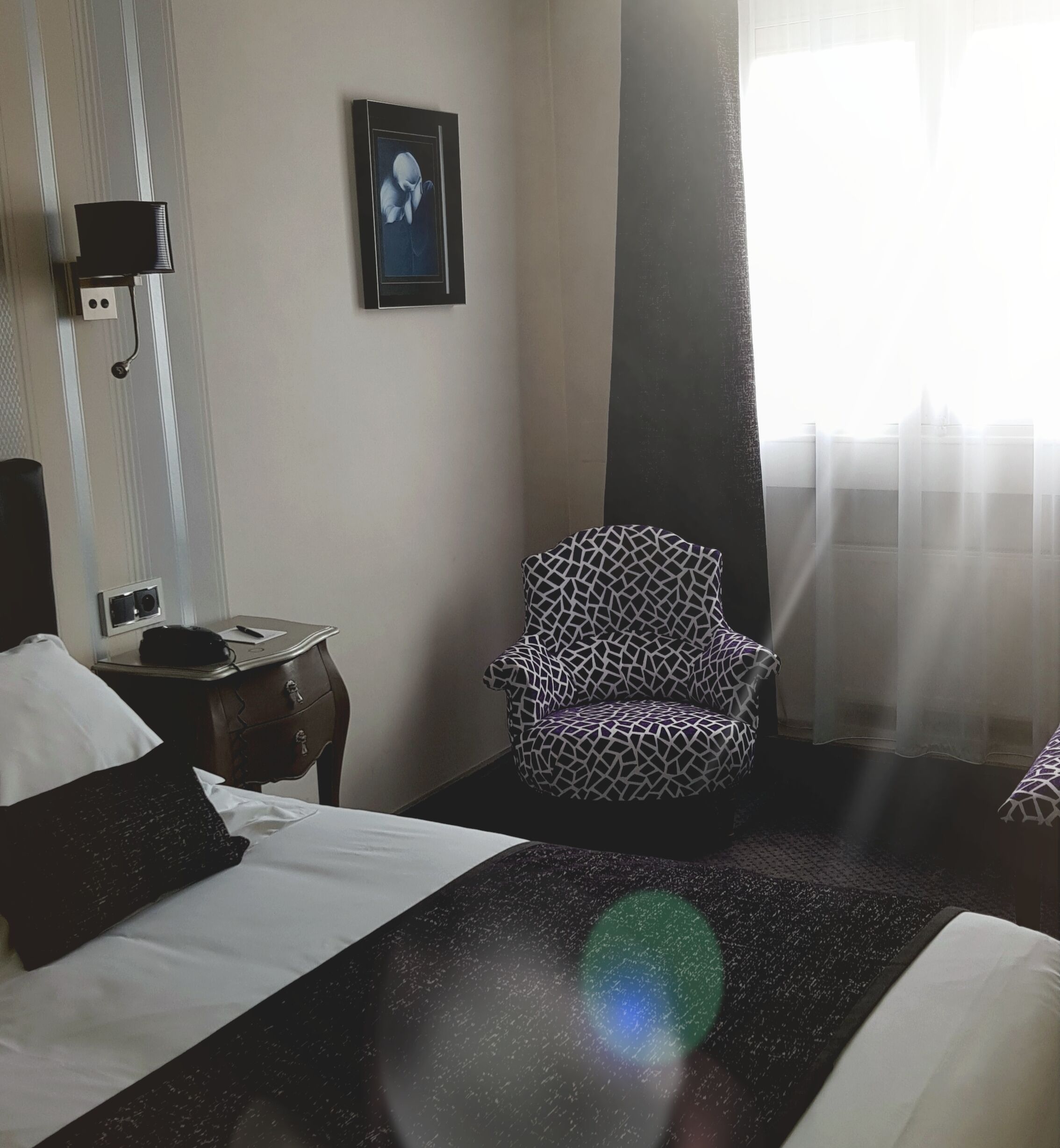 A hotel room with a bed, a patterned armchair, a nightstand, a wall-mounted lamp, and a framed picture.
