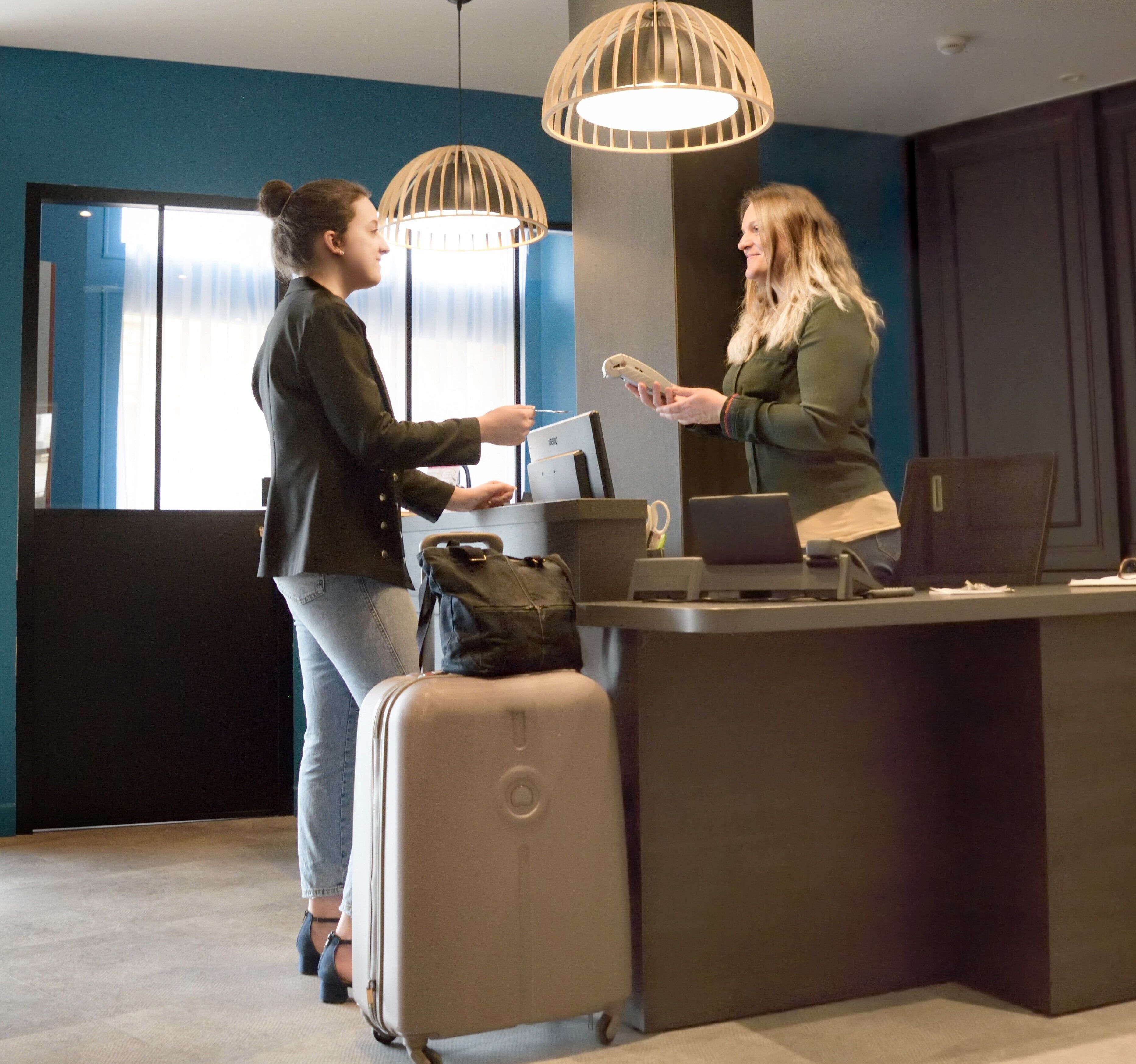 Due persone alla reception di un moderno hotel, con un'ampia vetrata, una scrivania con un portatile e una valigia.