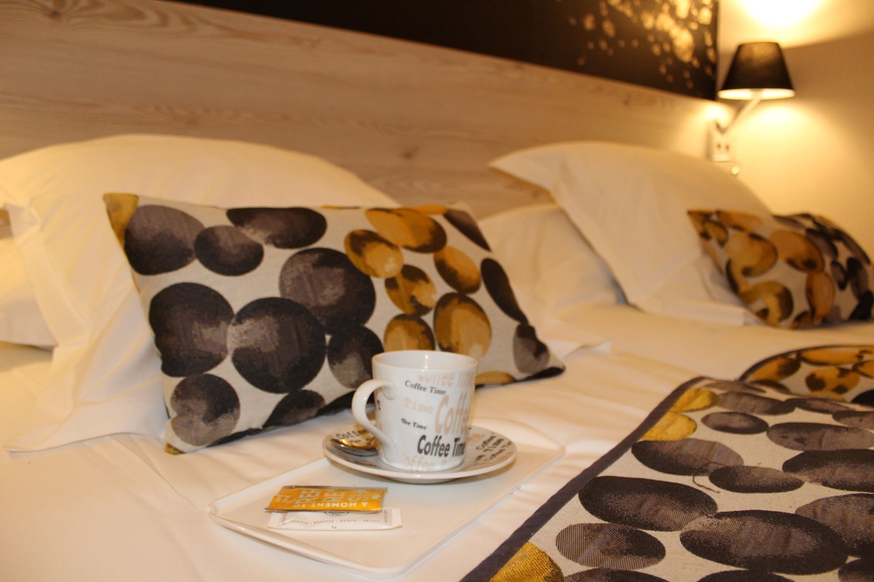 A hotel room with a bed featuring a coffee-themed pillow design, a cup of coffee, and a packet of coffee creamer.