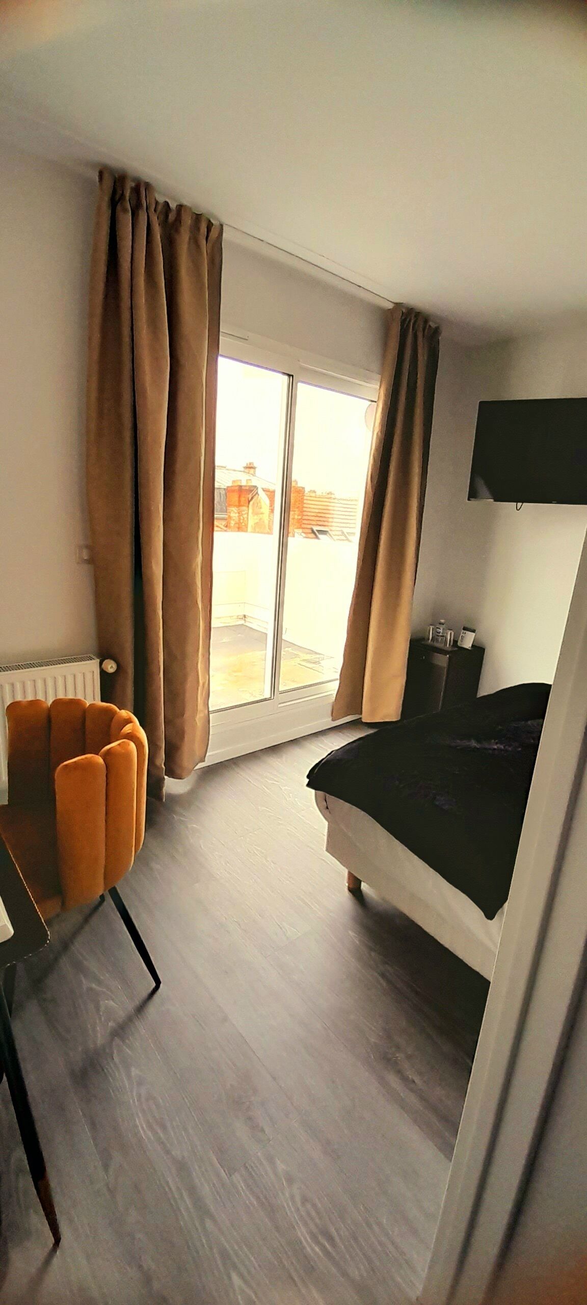 A small, modern room with a bed, a desk, and a chair. There is a window with curtains, a radiator, and a TV mounted on the wall.