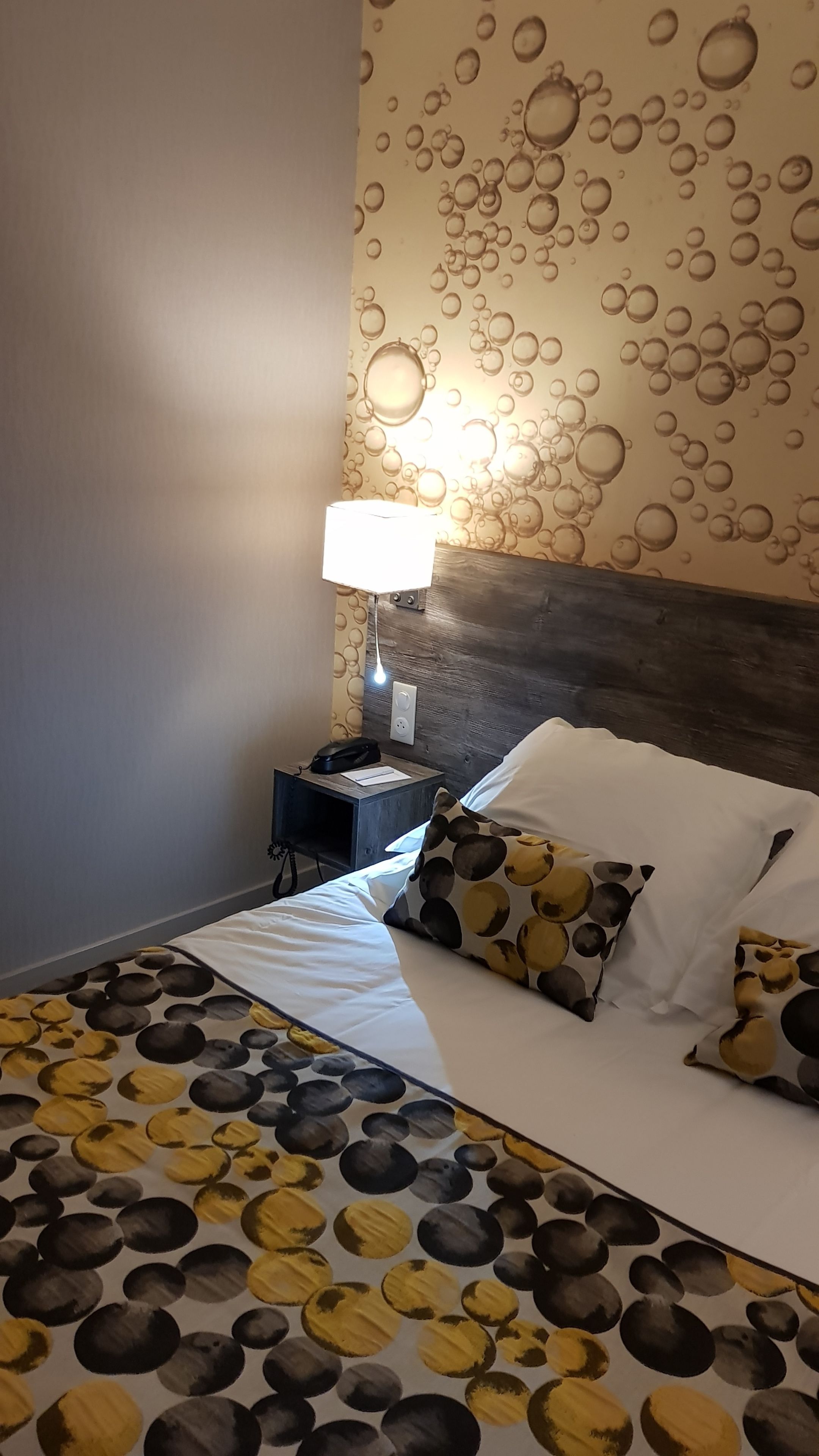A bedroom with a bed, pillows, a nightstand, a lamp, and a wall with bubble patterns.