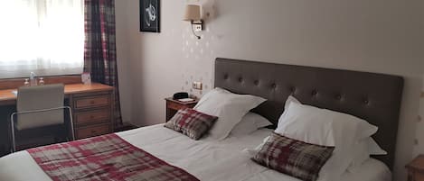 A neatly made bed with a plaid blanket and two pillows, a desk with a chair, a nightstand with a lamp, and a wall clock.