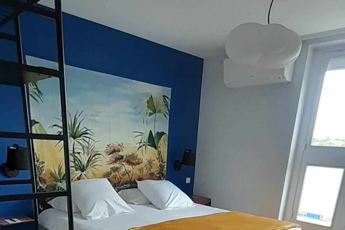A bedroom with a bed, a yellow chair, a painting on the wall, and a window.