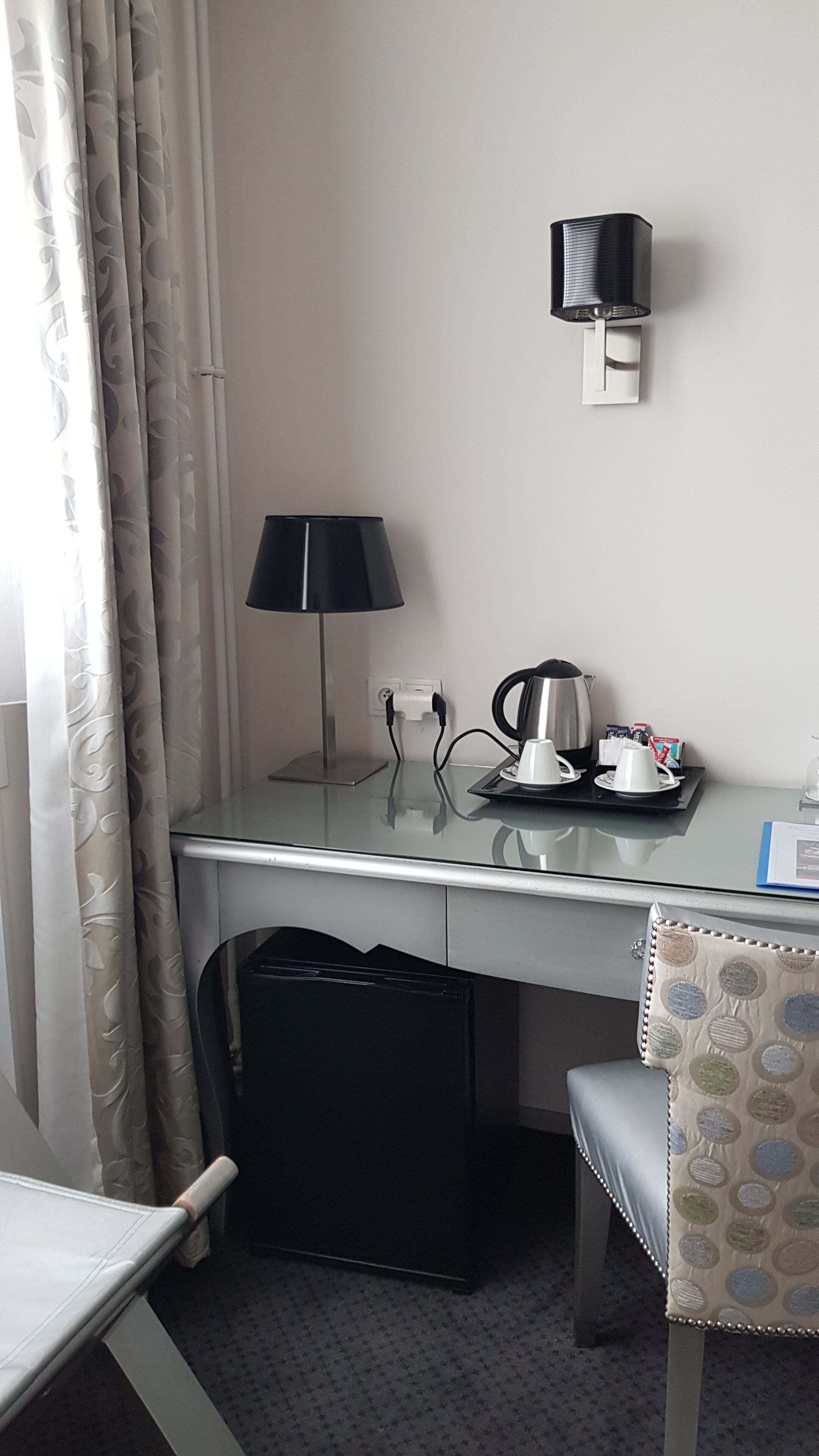 Habitació d'hotel amb escriptori de vidre, làmpada negra, un brollador d'aigua, tassa i cadira amb coixí estampat.