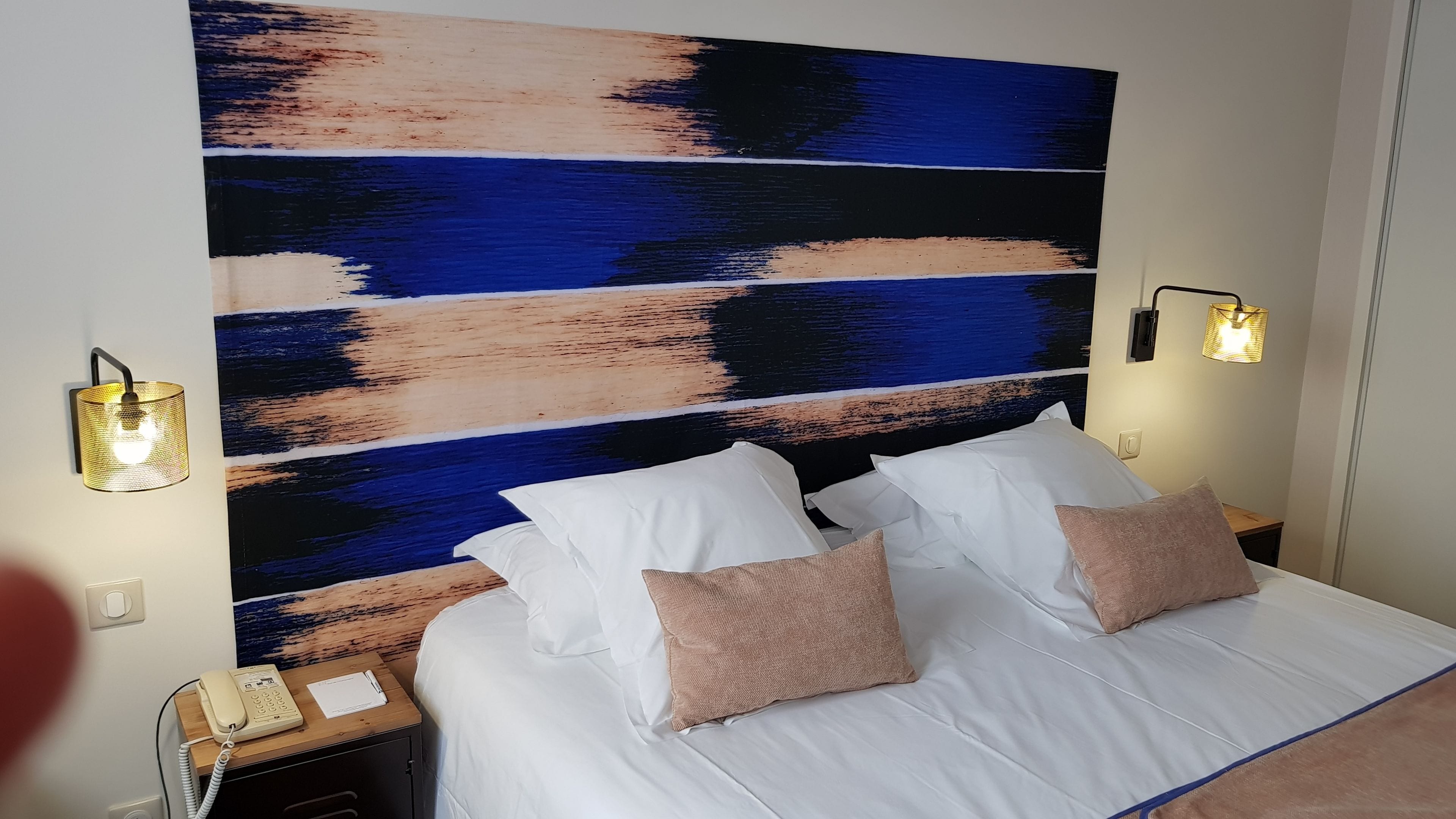 Una habitación de hotel con un cuadro abstracto grande en la pared, una cama con sábanas blancas y dos almohadas, una mesita de noche con una lámpara y un teléfono.