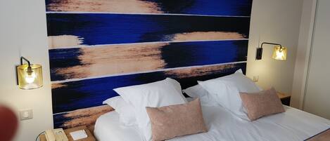 Una habitación de hotel con un cuadro abstracto grande en la pared, una cama con sábanas blancas y dos almohadas, una mesita de noche con una lámpara y un teléfono.