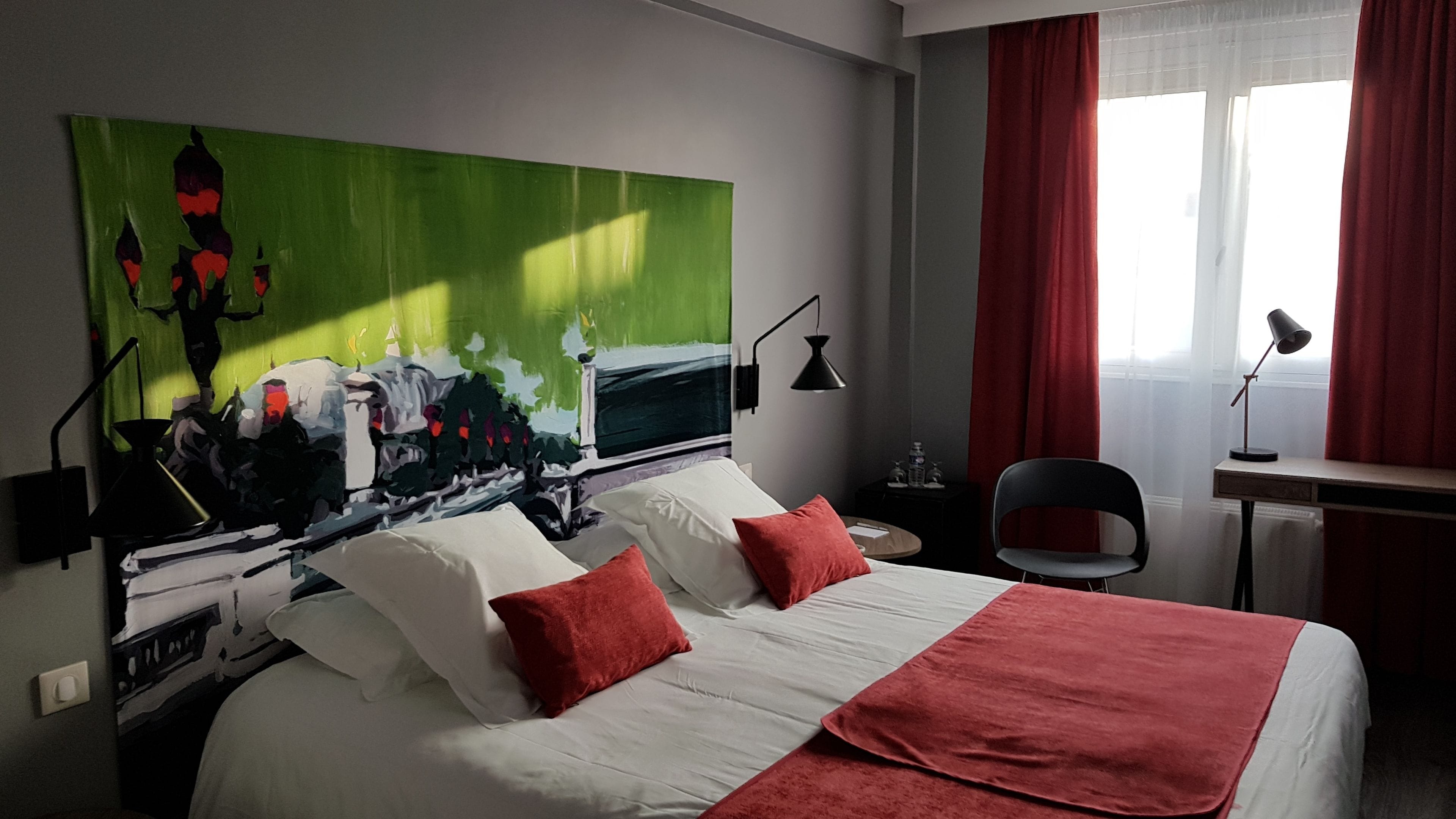 A hotel room with a large bed, red pillows, a green abstract painting, a desk with a chair, and a window with red curtains.