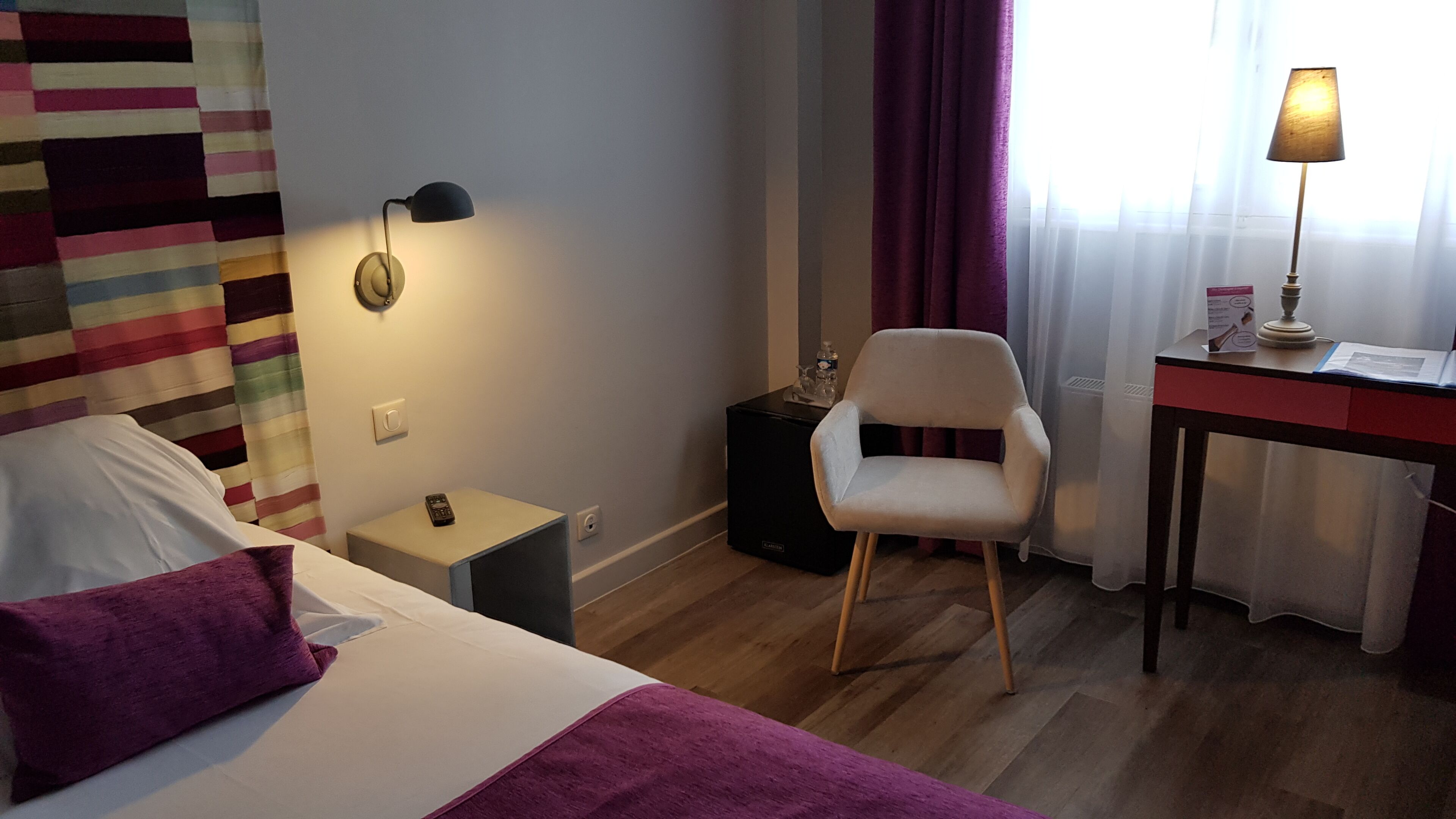 Une chambre d’hôtel comprenant un lit, une chaise, un bureau, une lampe, une horloge et une petite table avec une télécommande.