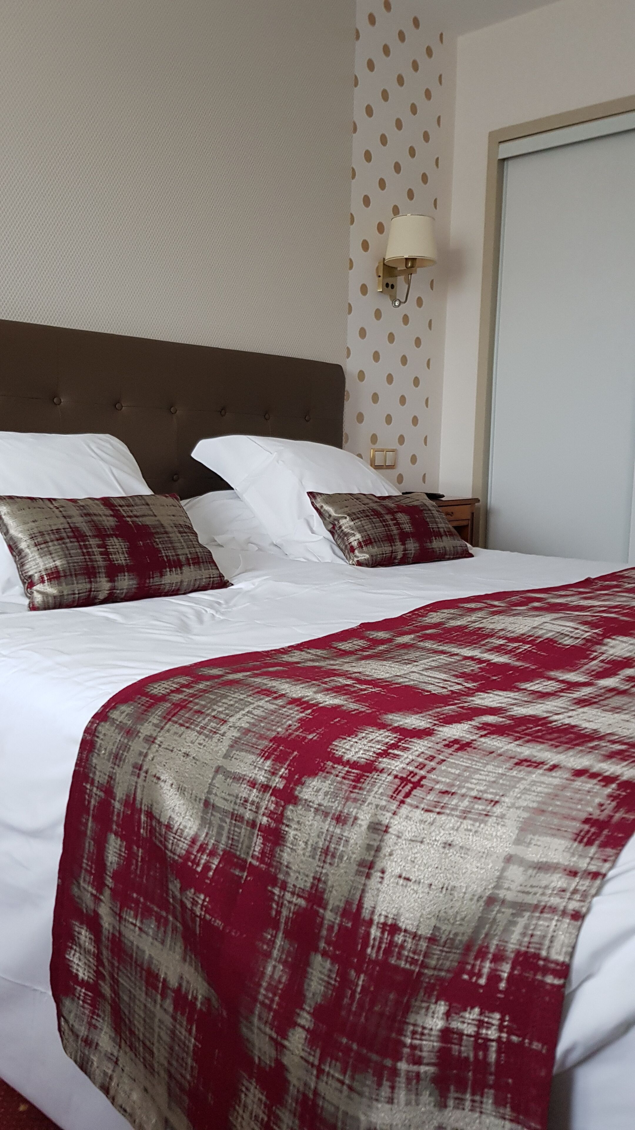 Una cama bien tendida con sábanas blancas y una colcha a cuadros rojos y blancos. Hay dos almohadas, una con un estampado rojo y blanco y otra con diseño a cuadros. La habitación tiene un cabecero marrón y una lámpara de pared.