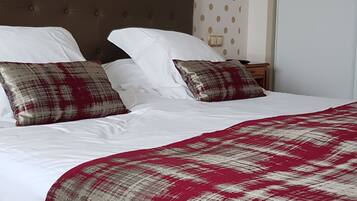 Una cama bien tendida con sábanas blancas y una colcha a cuadros rojos y blancos. Hay dos almohadas, una con un estampado rojo y blanco y otra con diseño a cuadros. La habitación tiene un cabecero marrón y una lámpara de pared.