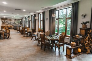 Un comedor amplio con mesas y sillas de madera, ventanas grandes con cortinas, y lámparas fijadas a la pared.