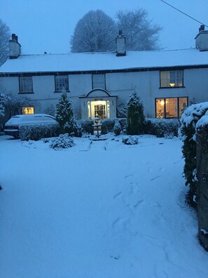 A snow-covered house with a well-lit entrance, a visible car, and a snow-covered garden. - Old School House (Ambleside)