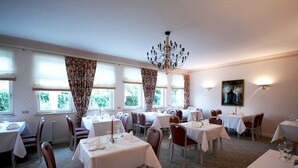 A dining area with multiple tables and chairs, large windows with curtains, and a chandelier. - Parkhotel (Nieheim)
