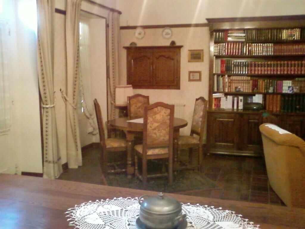 Een eethoek met een houten tafel, stoelen en een kanttafelkleed. Aan de rechterkant staat een boekenplank vol boeken.