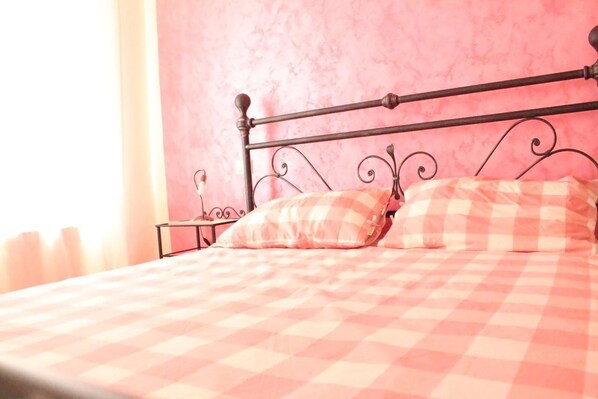 A bed with a checkered bedspread and two pillows, a nightstand with a lamp, and a pink wall. - L'Iris B&B (Lucca)