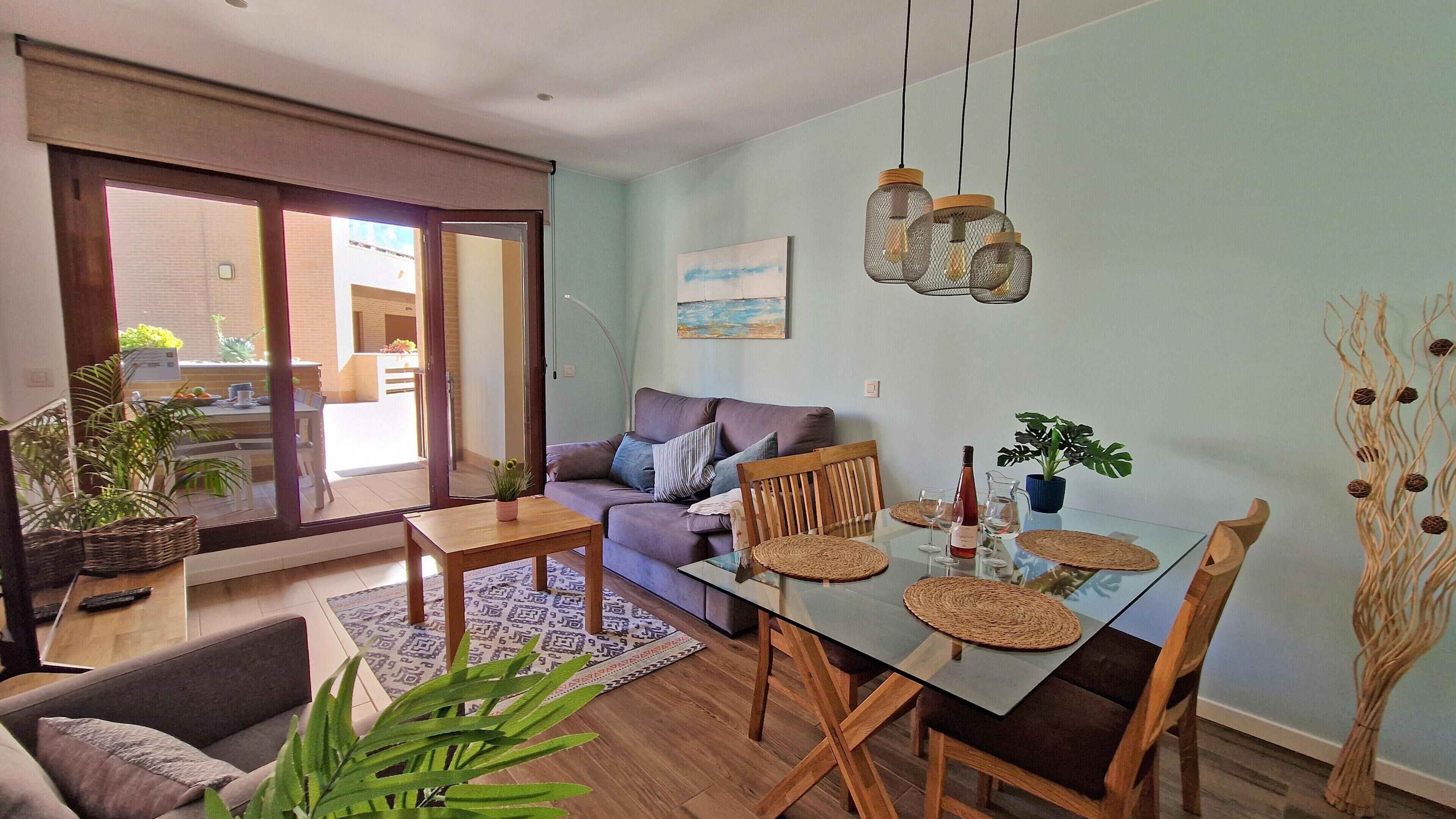 A modern living room with a glass dining table, wooden chairs, a sofa, and a large window with a view of the outside.