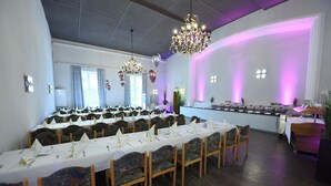 Een feestzaal met lange tafels gedekt voor een diner, houten stoelen, een kroonluchter en een podium met paars licht.