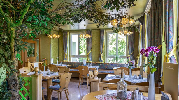 Een restaurant met houten tafels en stoelen, grote ramen en hangende lampen.