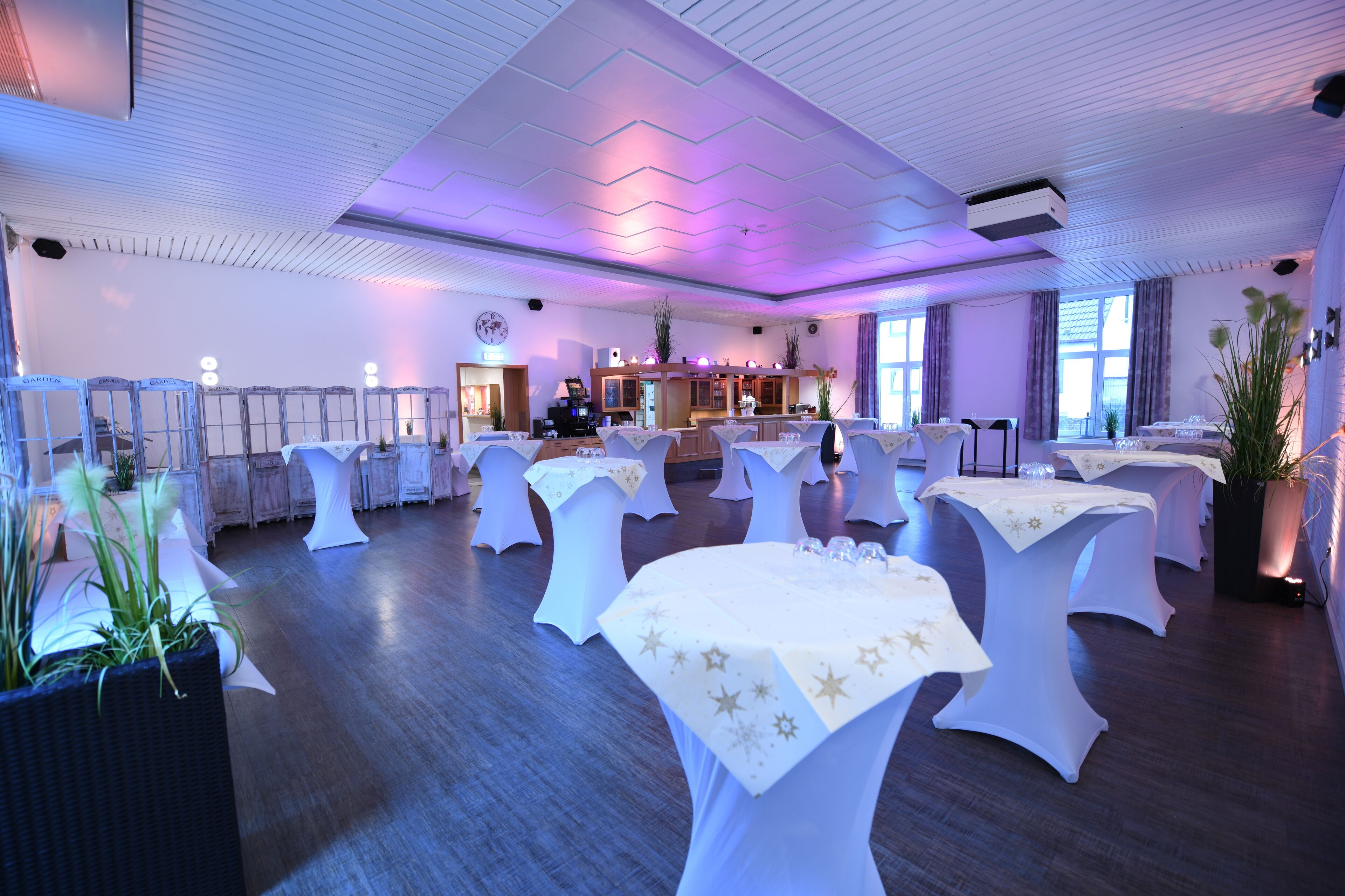 Un amplio salón de banquetes con varias mesas redondas cubiertas con manteles blancos, una zona de bar y grandes ventanales que permiten la entrada de luz natural.