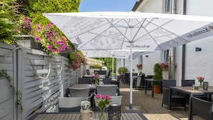 Een terras buiten met rieten meubels, witte parasols en plantenpotten.