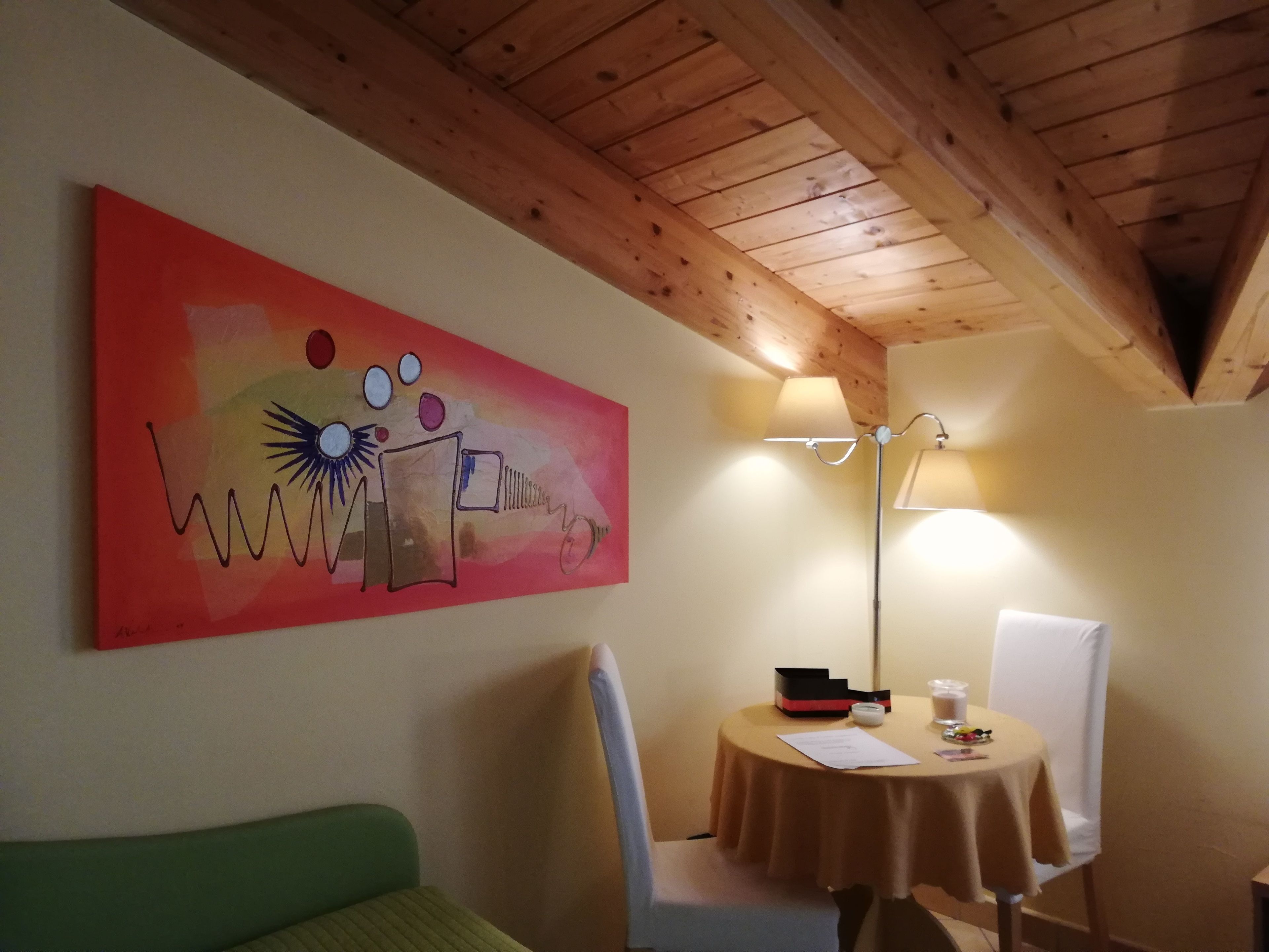 A room with a wooden ceiling, a green sofa, a table with a lamp, and a colorful abstract painting on the wall.