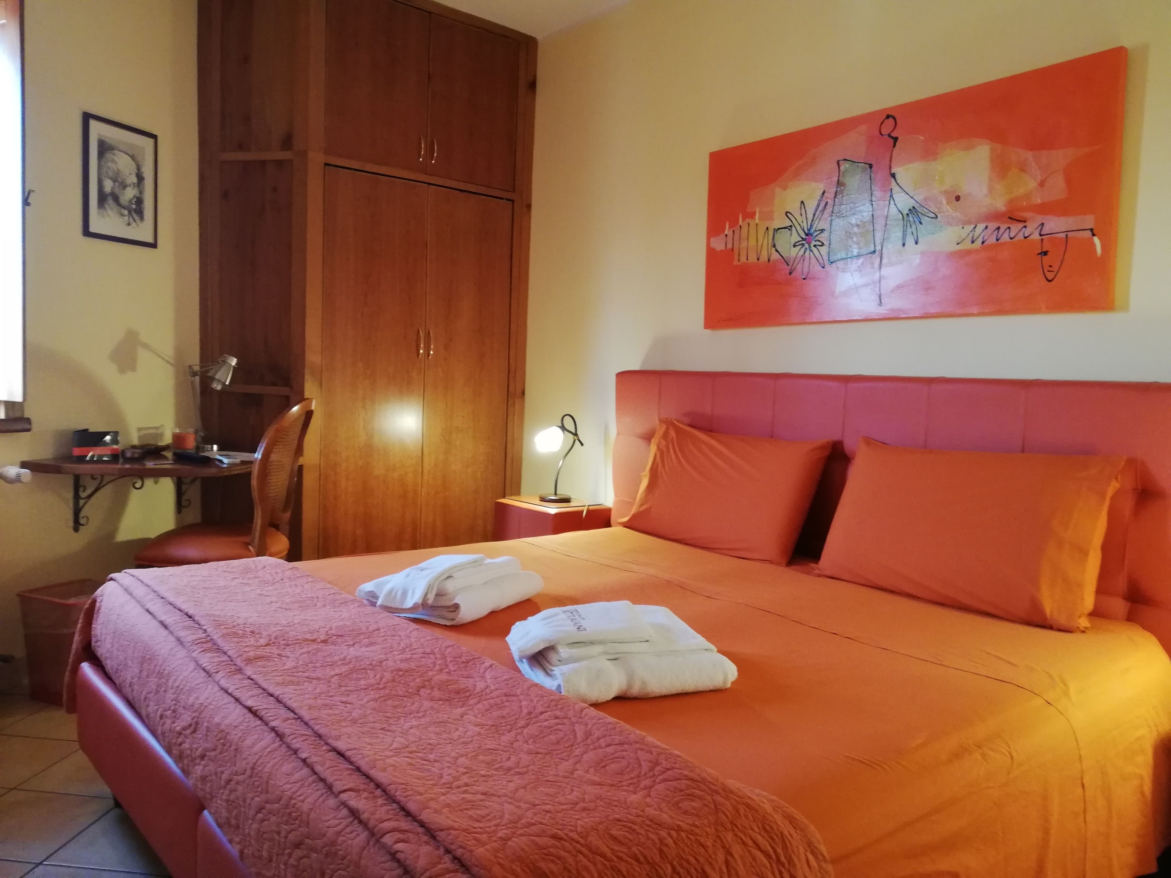 A hotel room with a large bed, orange bedding, a wooden wardrobe, a desk with a lamp, and a painting on the wall.