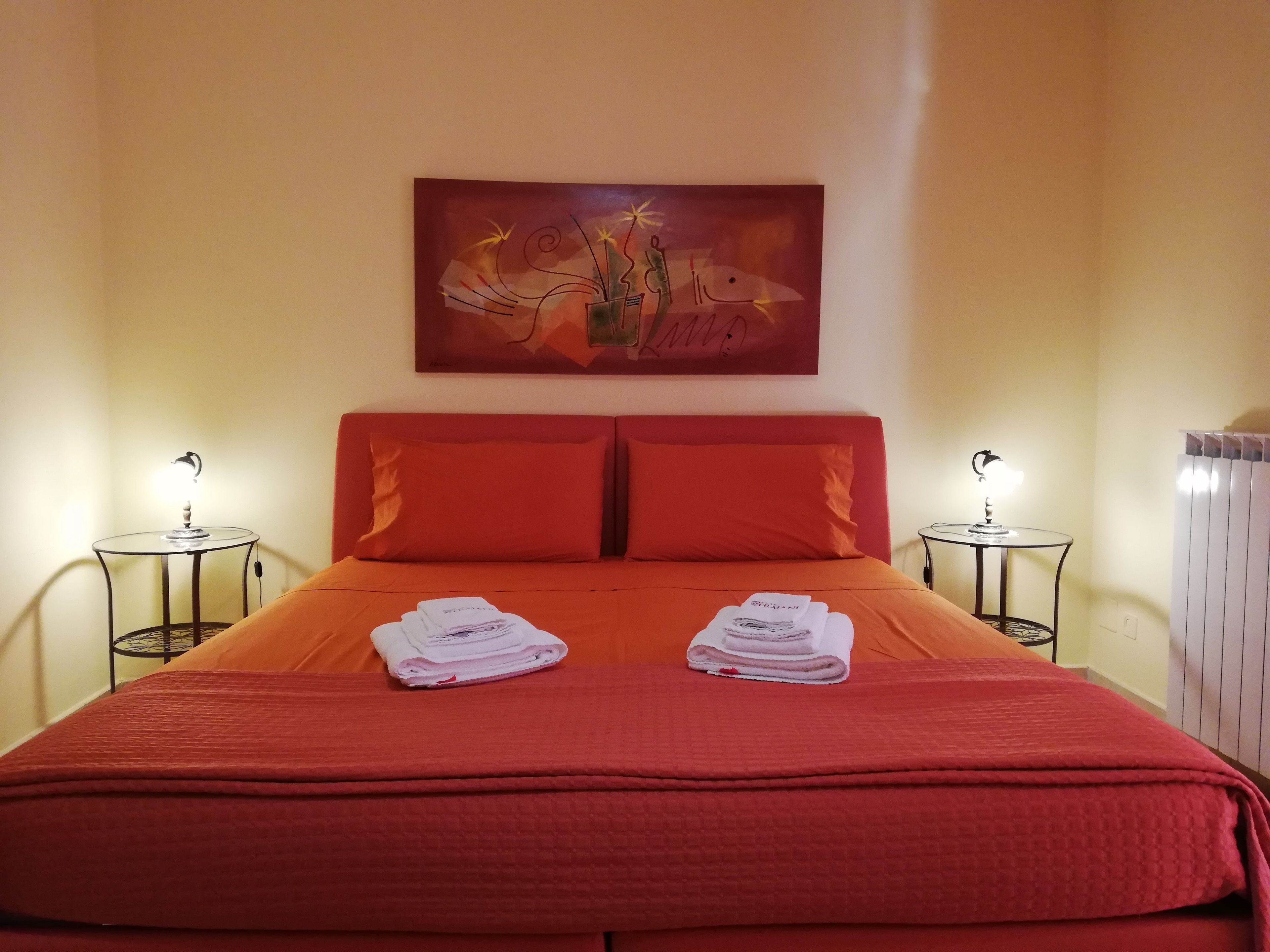 A hotel room with a red bed, two bedside tables with lamps, a painting on the wall, and a radiator.