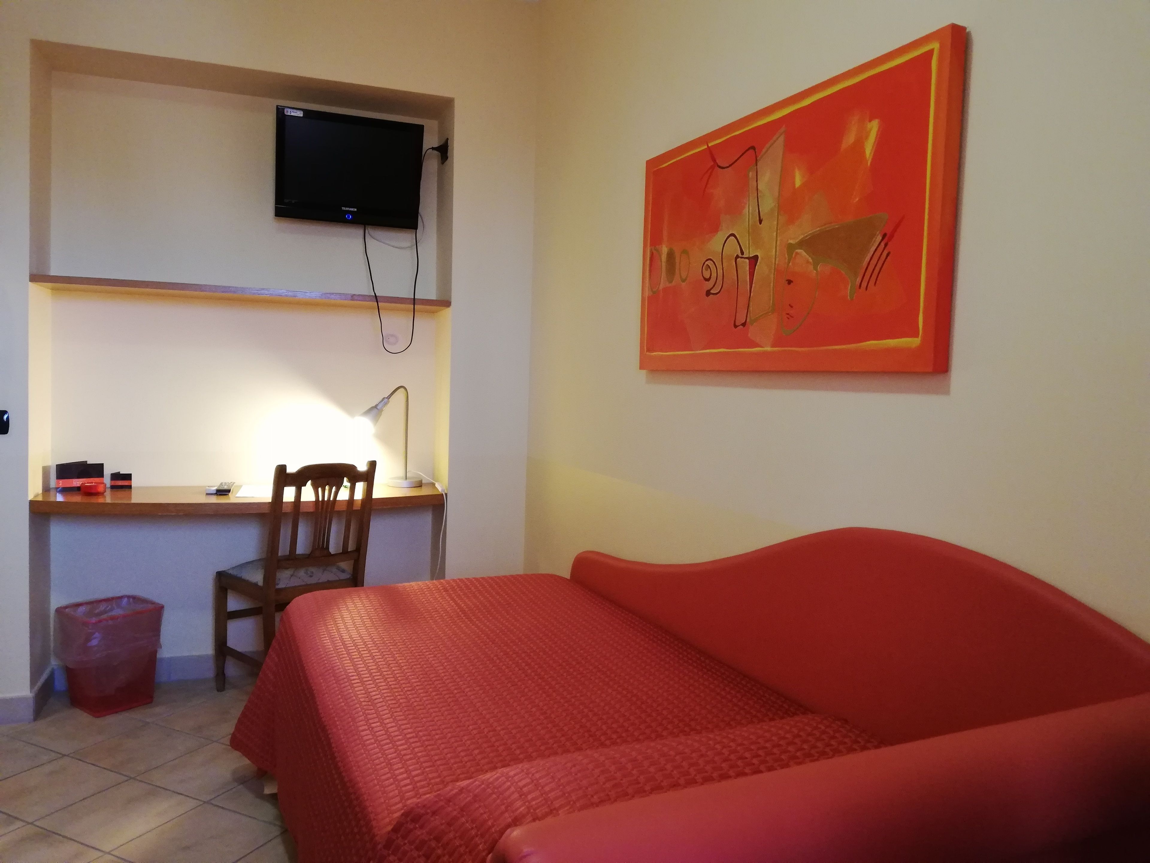 A room with a bed, a desk, a chair, a TV mounted on the wall, and a painting.