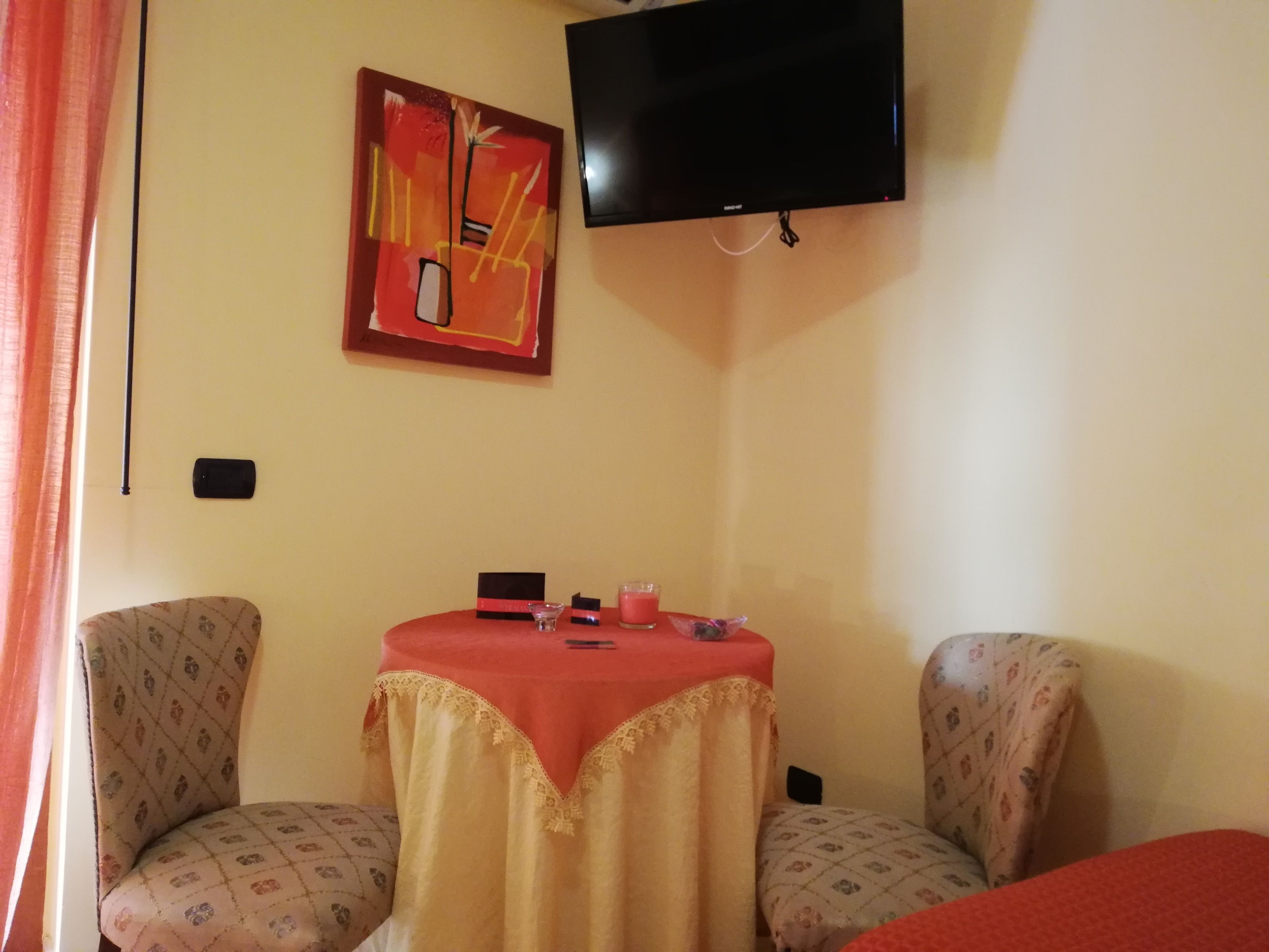 Una habitación con dos sillones estampados, una mesita con mantel rojo, un televisor de pantalla plana en la pared y un cuadro encima del televisor.