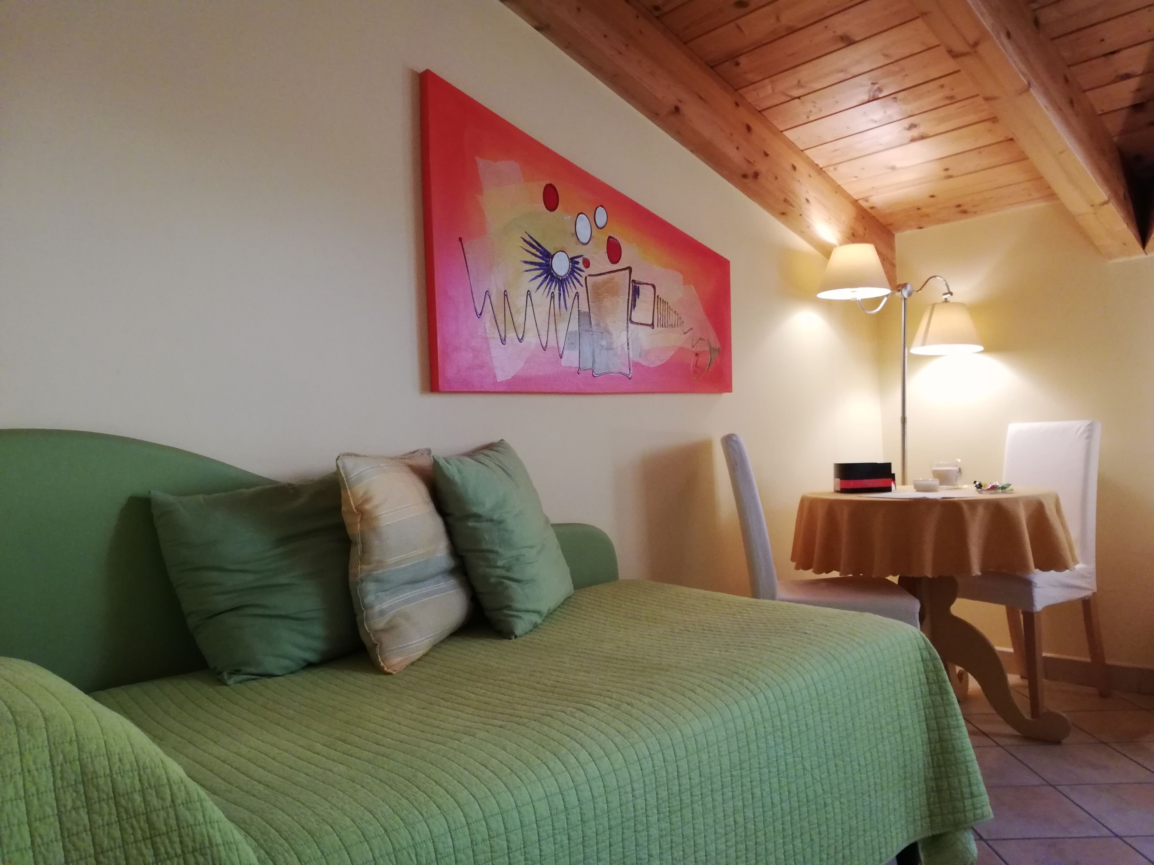 A room with a green bed, a painting on the wall, a table with a lamp, and a chair.