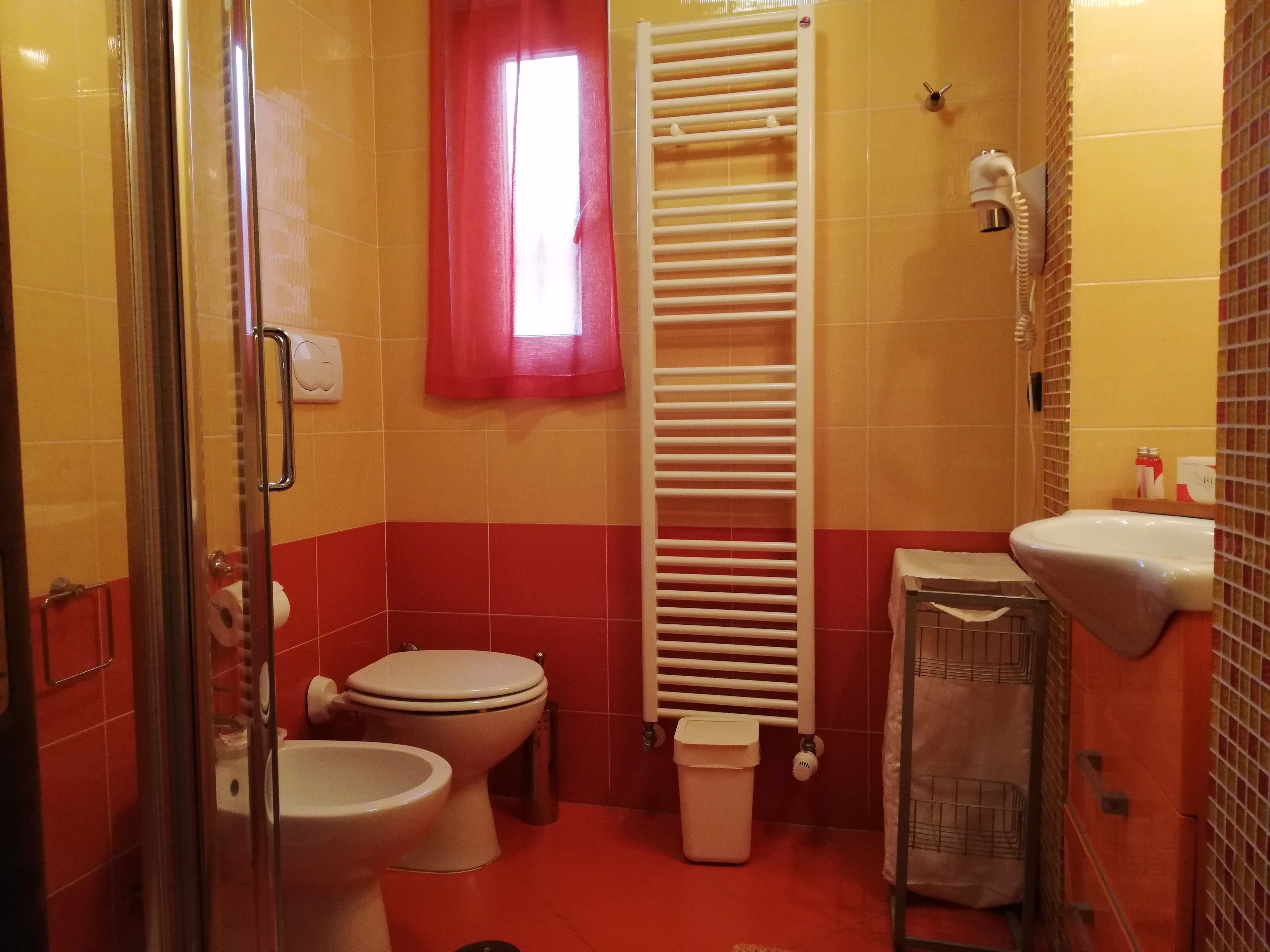 A bathroom with a glass shower enclosure, a toilet, a bidet, a white sink, a towel rack, a red-framed window, and a white towel rack.
