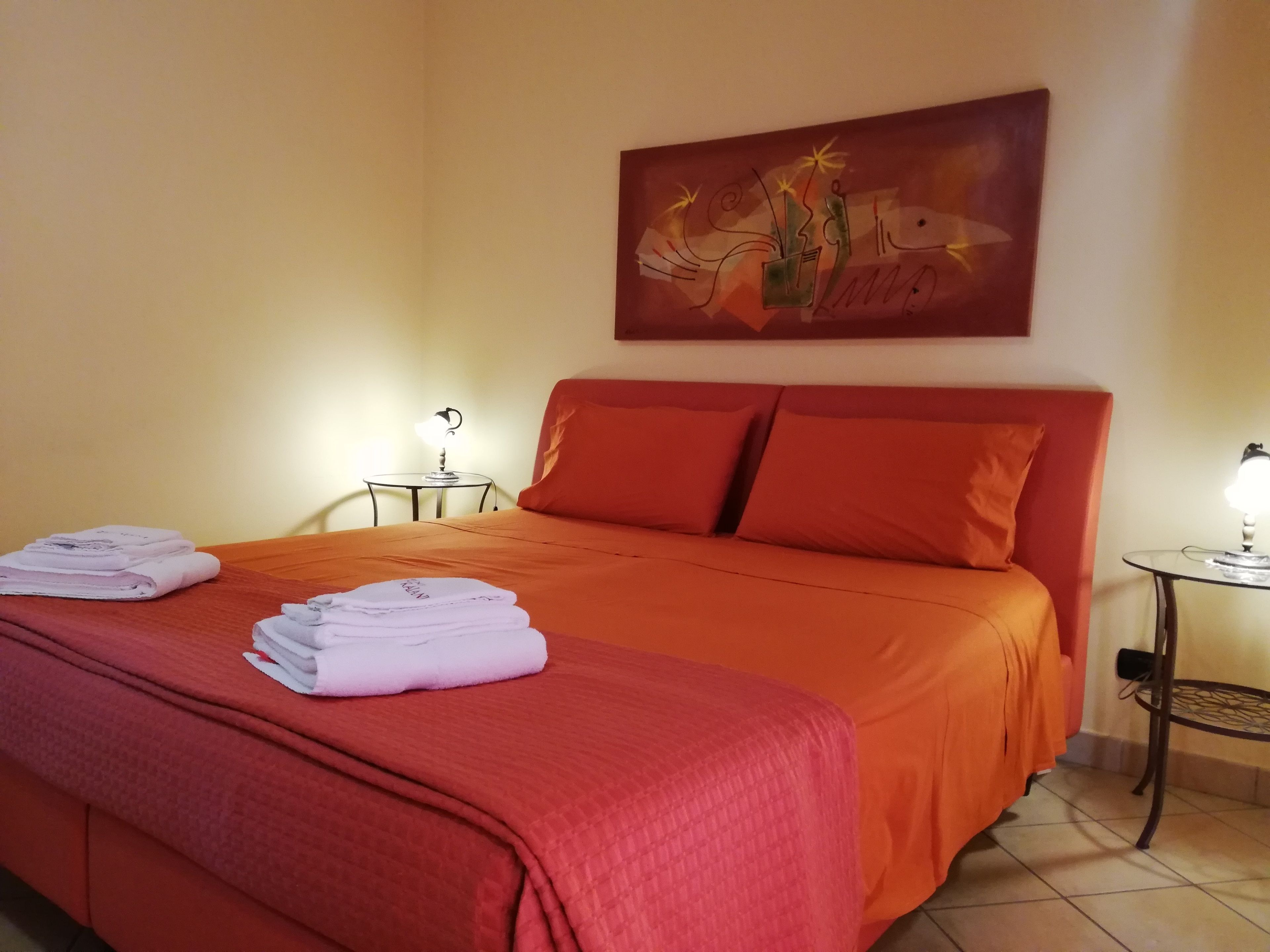 Cama bem arrumada com roupa de cama vermelha e toalhas dobradas ao lado. Há uma pequena mesa com abajur ao lado da cama e outra mesa com abajur no canto.