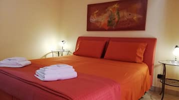 Cama bem arrumada com roupa de cama vermelha e toalhas dobradas ao lado. Há uma pequena mesa com abajur ao lado da cama e outra mesa com abajur no canto.