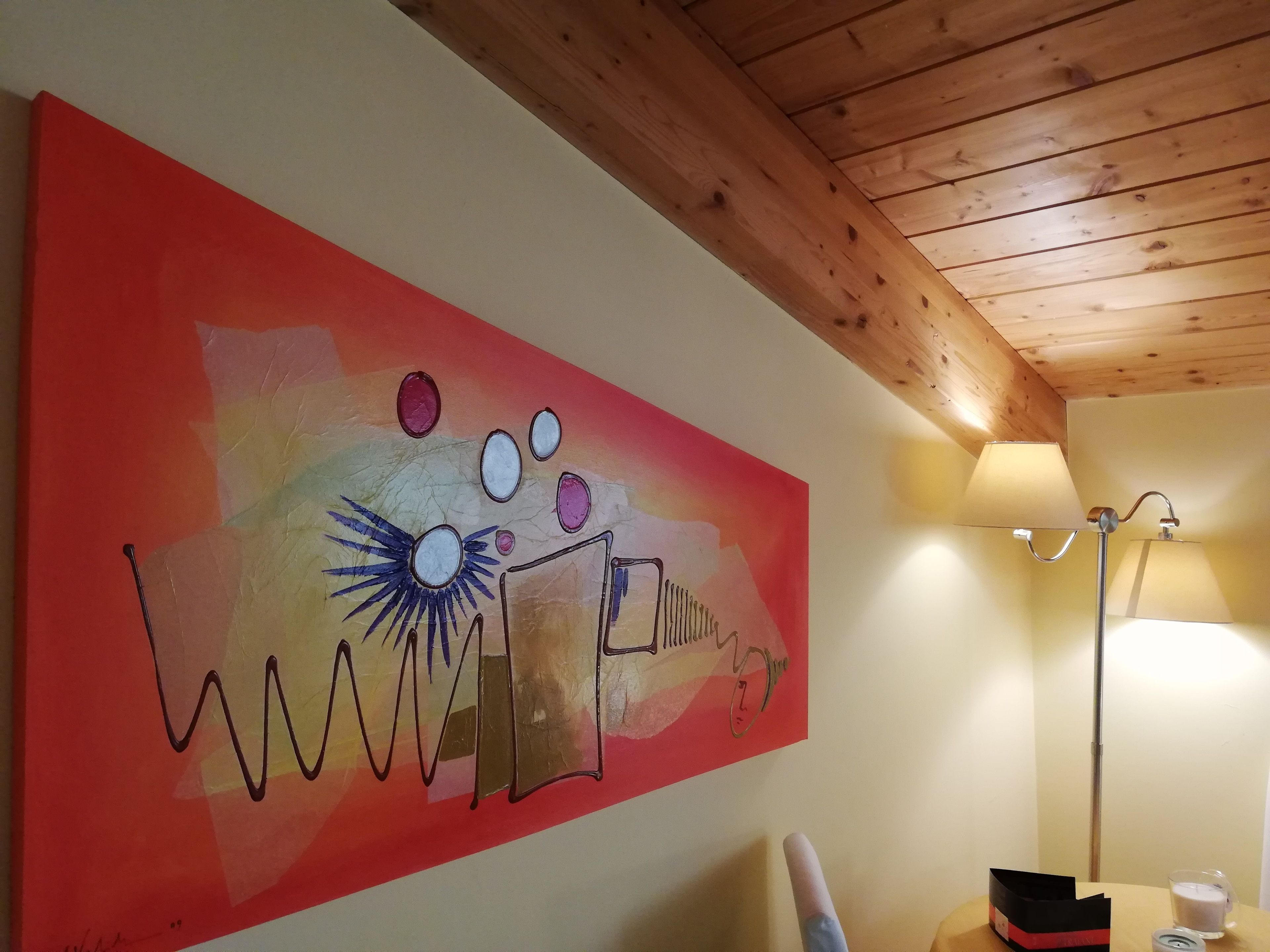 A room with a wooden ceiling, a painting on the wall, a lamp, and a table with a candle and a box.