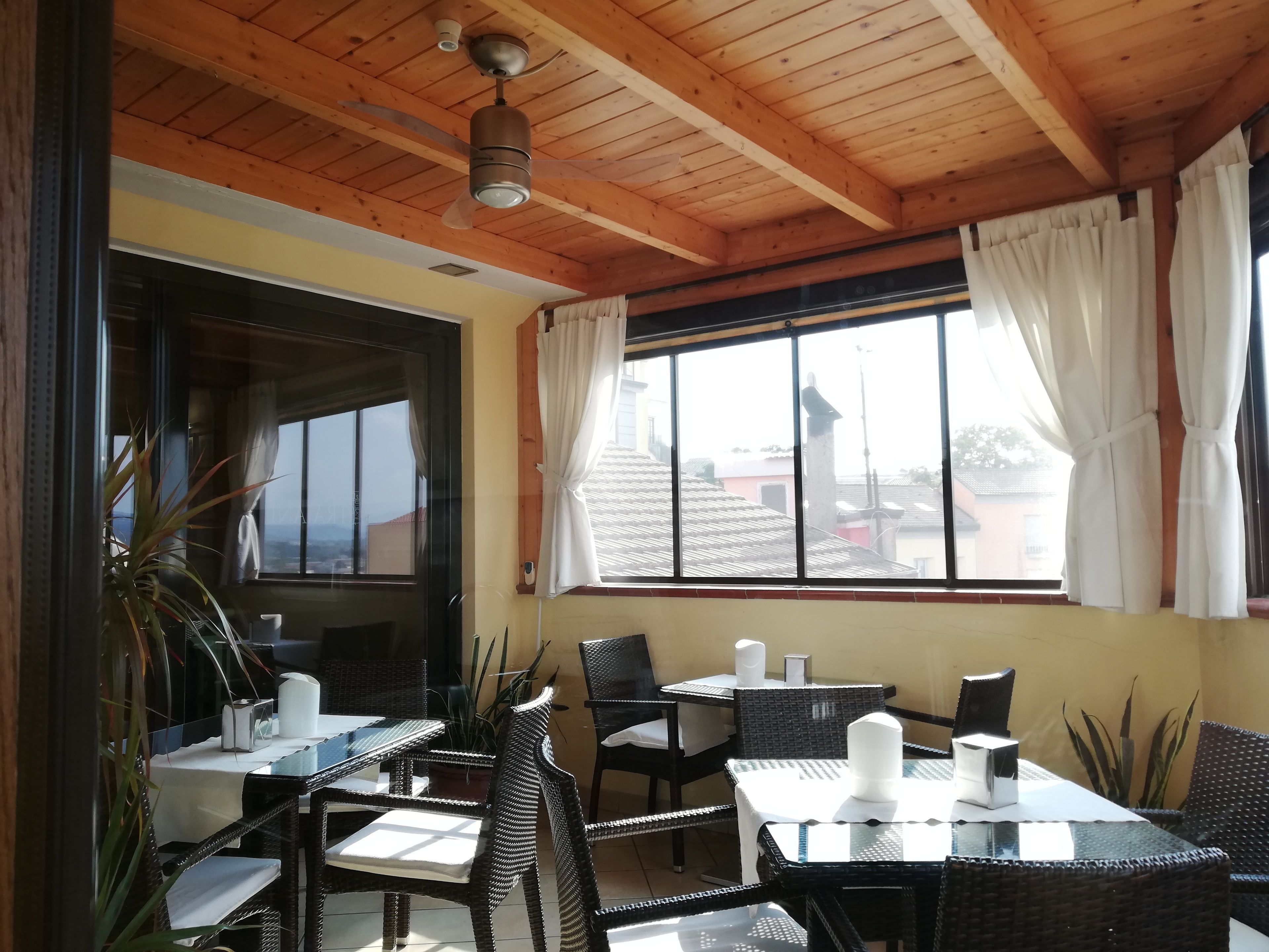 Un patio cubierto con mobiliario de mimbre, un ventilador de techo y vistas a edificios a través de amplios ventanales.