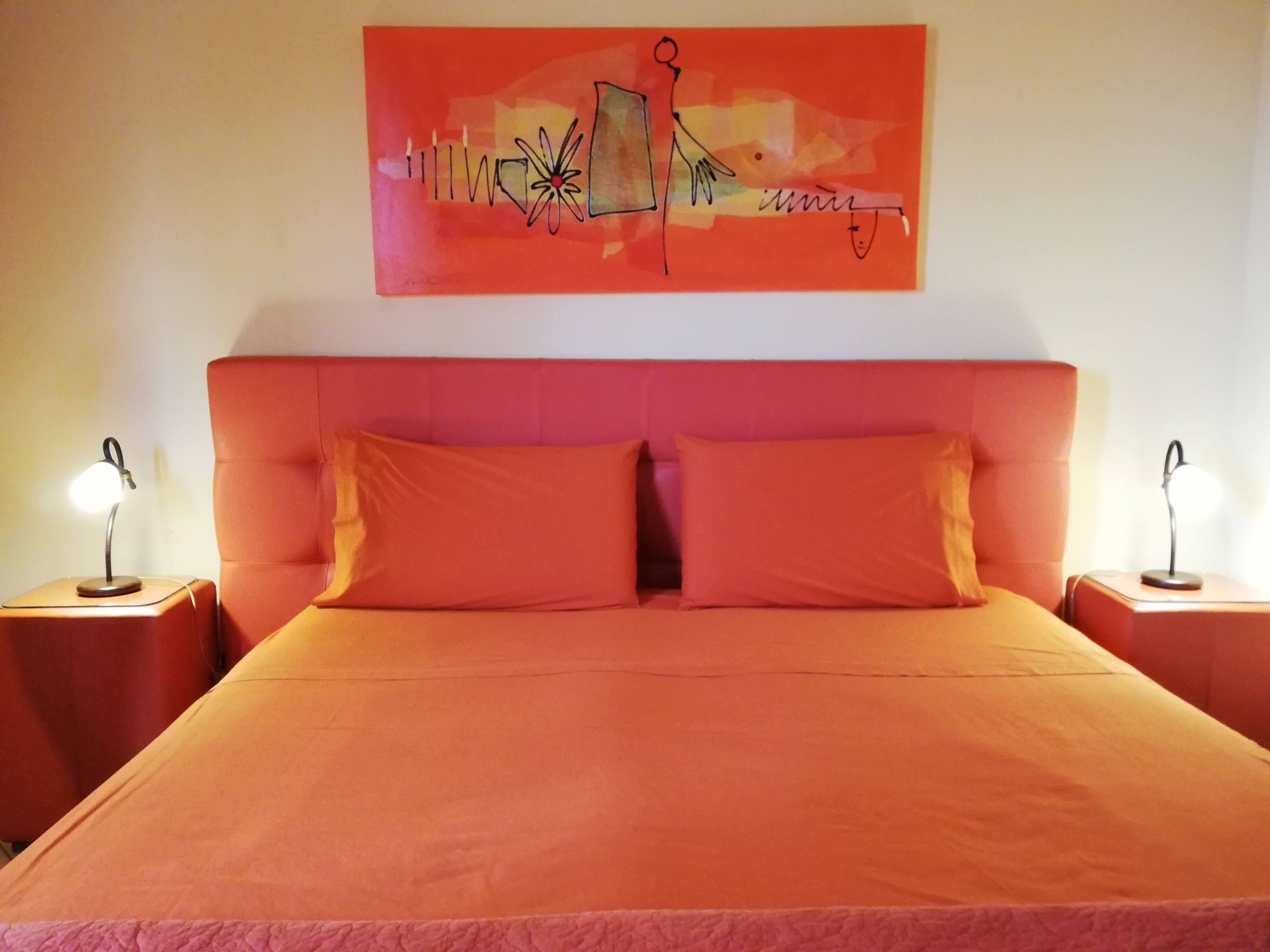 A bed with an orange headboard and two pillows, flanked by nightstands with lamps, in a room with a red abstract painting on the wall.