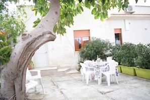 A patio with a white table and chairs, surrounded by potted plants and a tree. - Kitesurf lo Stagnone (Marsala)