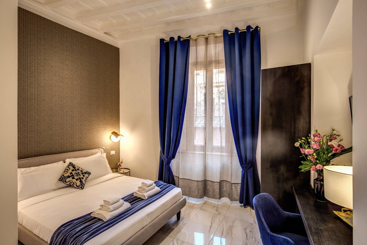 Una camera d'albergo con un letto, un comodino, una scrivania con una lampada e una sedia.