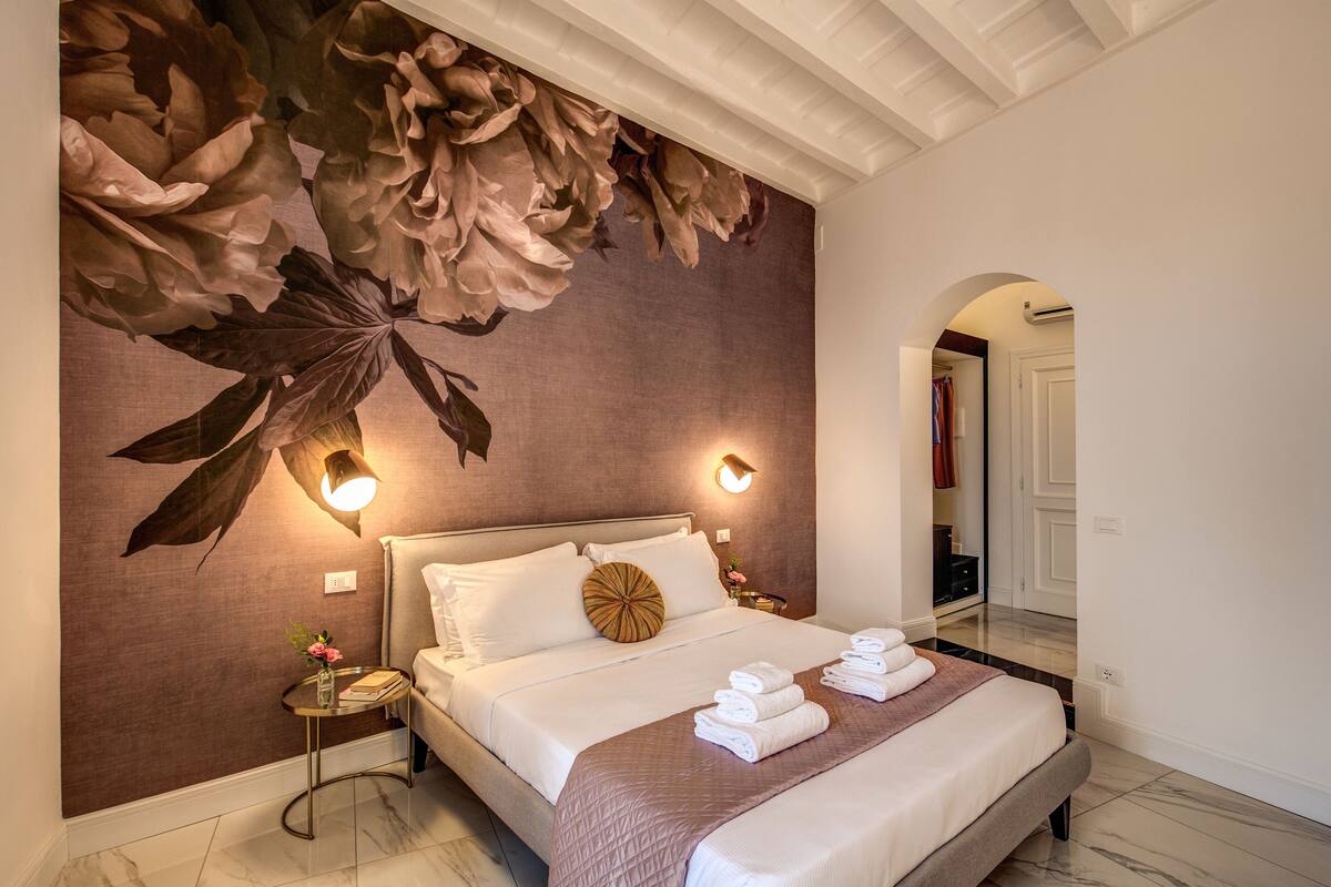 Una camera d'albergo con un letto grande, una sveglia rotonda a parete, un comodino e un ampio murale floreale.