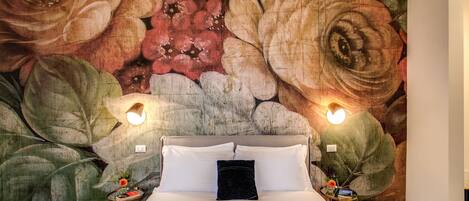 A bedroom with a large floral mural on the wall, a bed with white linens, two bedside tables with lamps, and a black throw pillow.