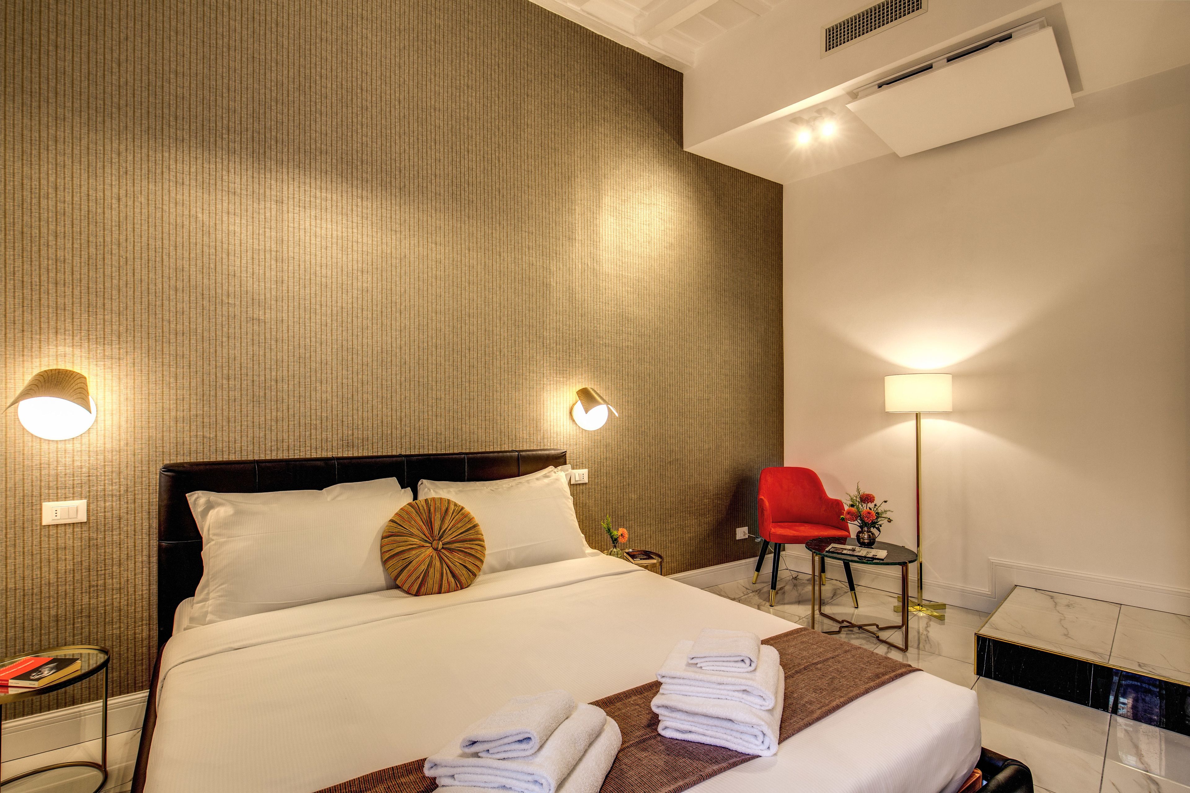 Quarto de hotel com uma cama grande, duas luminárias de cabeceira, uma cadeira vermelha, uma mesinha com abajur e ar-condicionado fixado na parede.