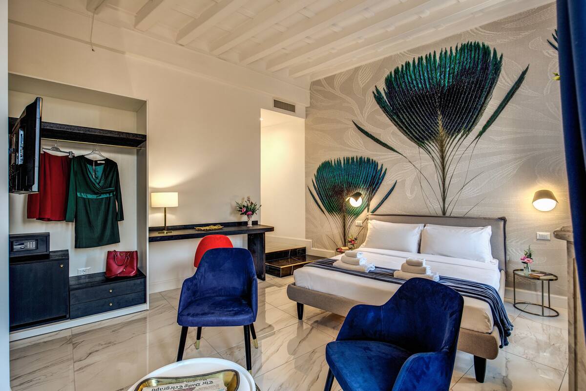 Camera d'albergo con un letto, due poltrone blu, una scrivania con sedia, un armadio e un muro decorato con un motivo a piuma di pavone.