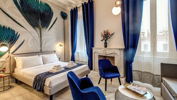 A modern bedroom with a large bed, a fireplace, and a seating area with blue chairs. - Little Queen Pantheon Residence (Rome)