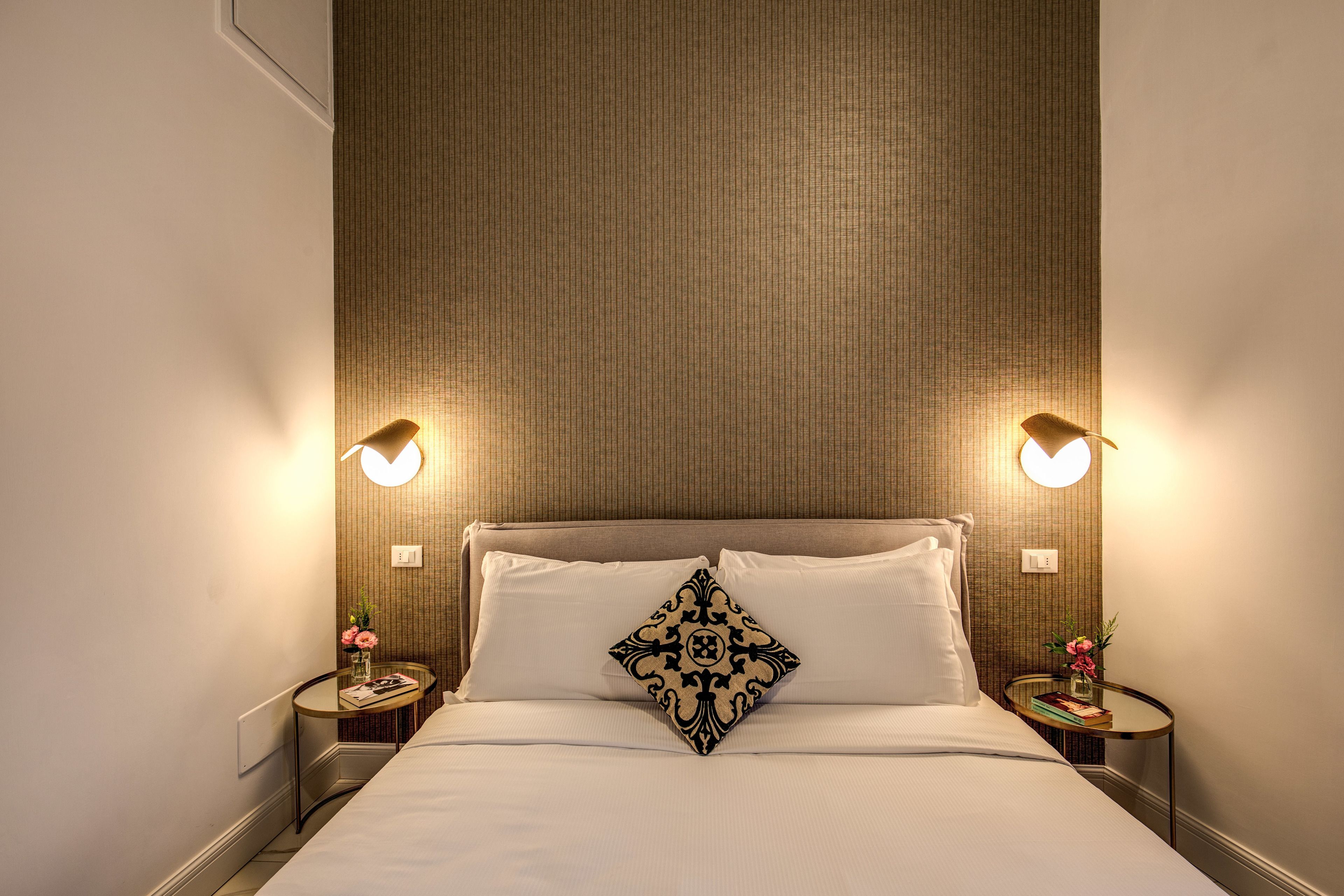 Una cama bien hecha con un cojín decorativo, dos mesitas de noche con lámparas, y una pared con líneas verticales.