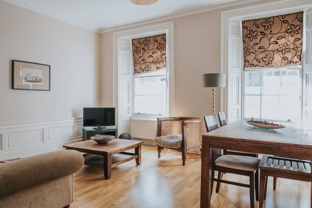 Ein Wohnzimmer mit Fernseher, einem Esstisch mit Stühlen, einer Couch und einem Fenster mit gemusterten Vorhängen.
