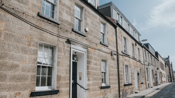 Une rangée de bâtiments traditionnels en pierre, dotés de fenêtres et de portes de style uniforme, une rue pavée et un ciel dégagé.