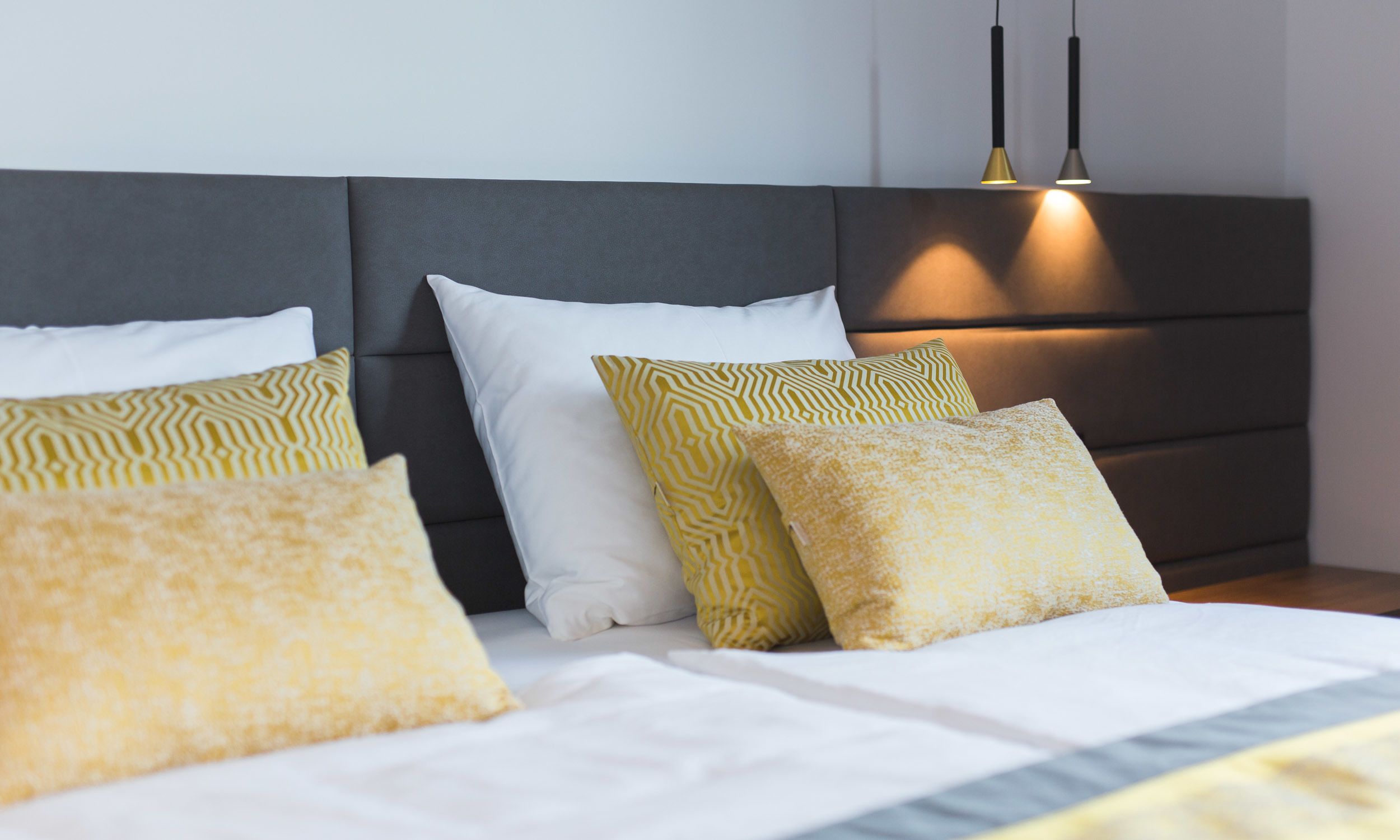 A neatly made bed with white and patterned pillows, a dark headboard, and two pendant lights.