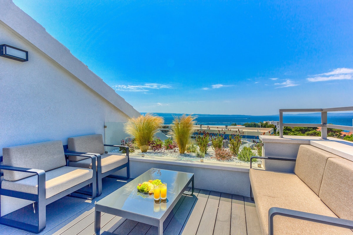 A rooftop terrace with outdoor seating, a table with fruit and drinks, and a view of the sea.