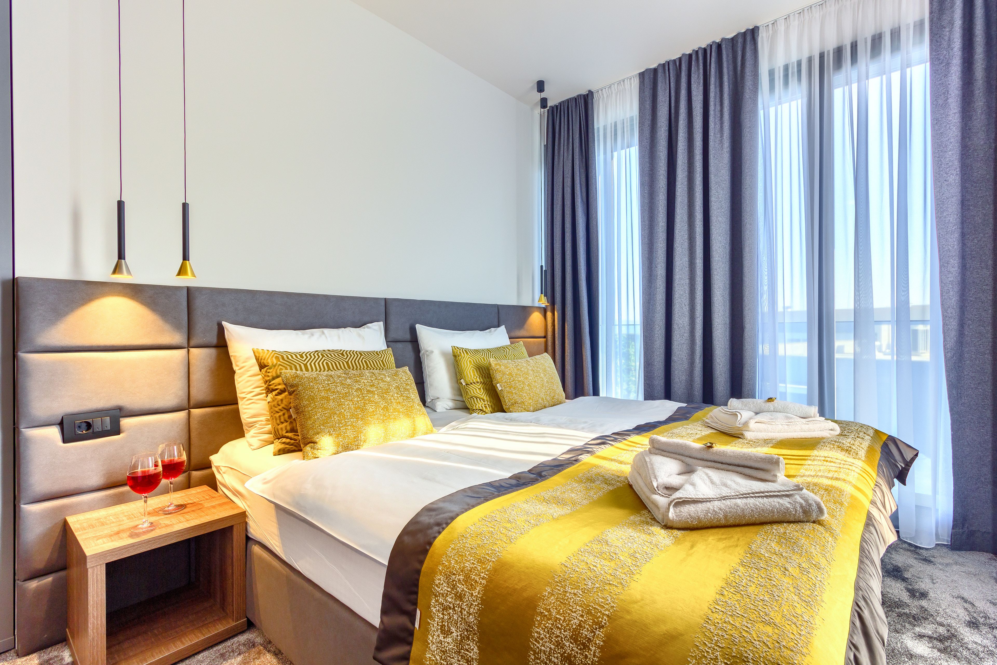 Une chambre d’hôtel avec un grand lit, une table de chevet sur laquelle reposent deux verres à vin, et une vue sur la ville à travers de fines rideaux.