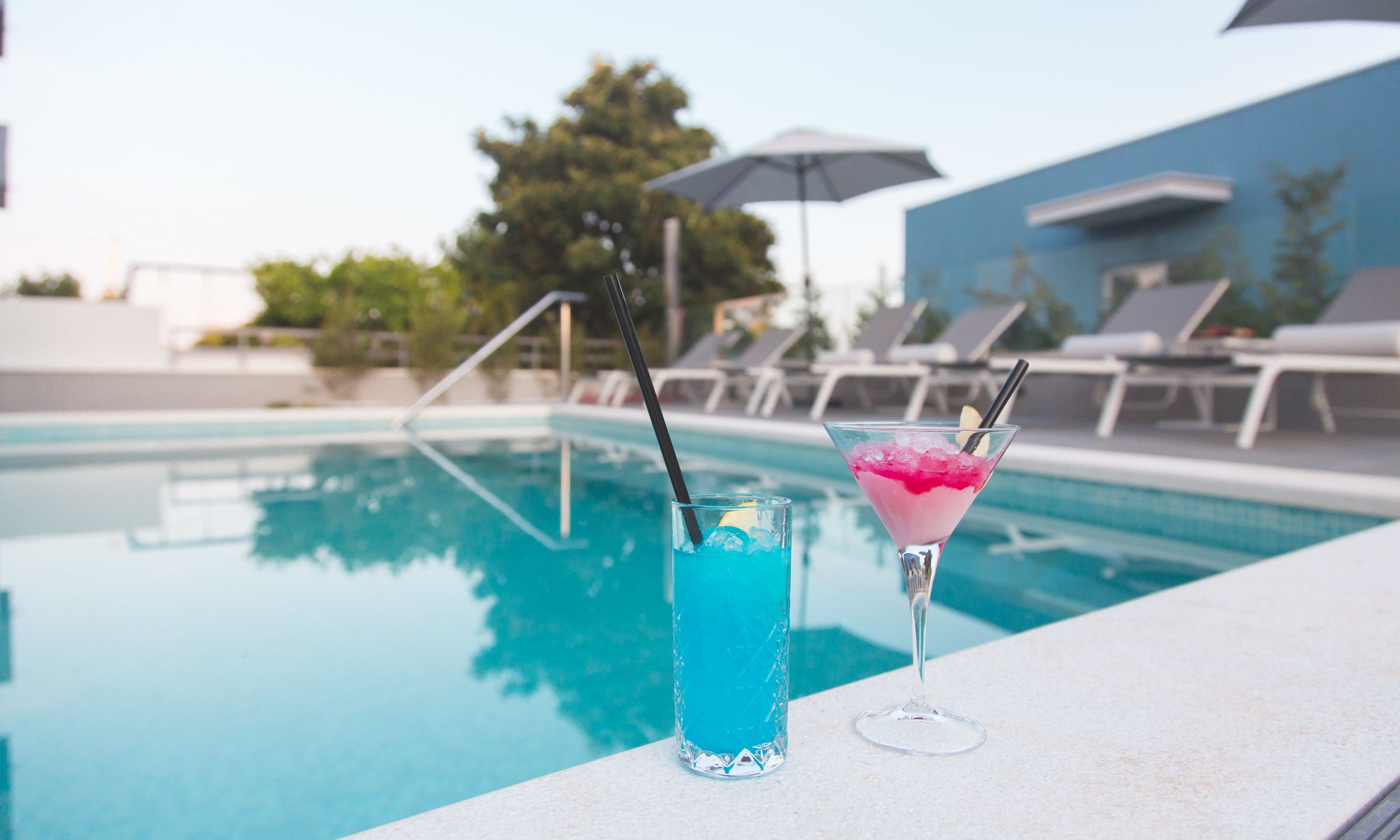 A poolside setting with two drinks, one in a tall glass and one in a martini glass, with a pool and lounge chairs in the background.
